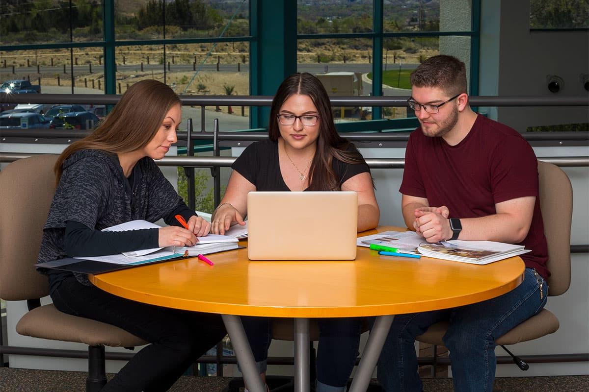 三个学生坐在一张桌子上，用一台笔记本电脑